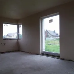 Surowe wnętrze pokoju z oknami, ściany po nałożeniu tynku maszynowego, widoczne wystające rury instalacji przy podłodze, posadzka betonowa przed wylewką.