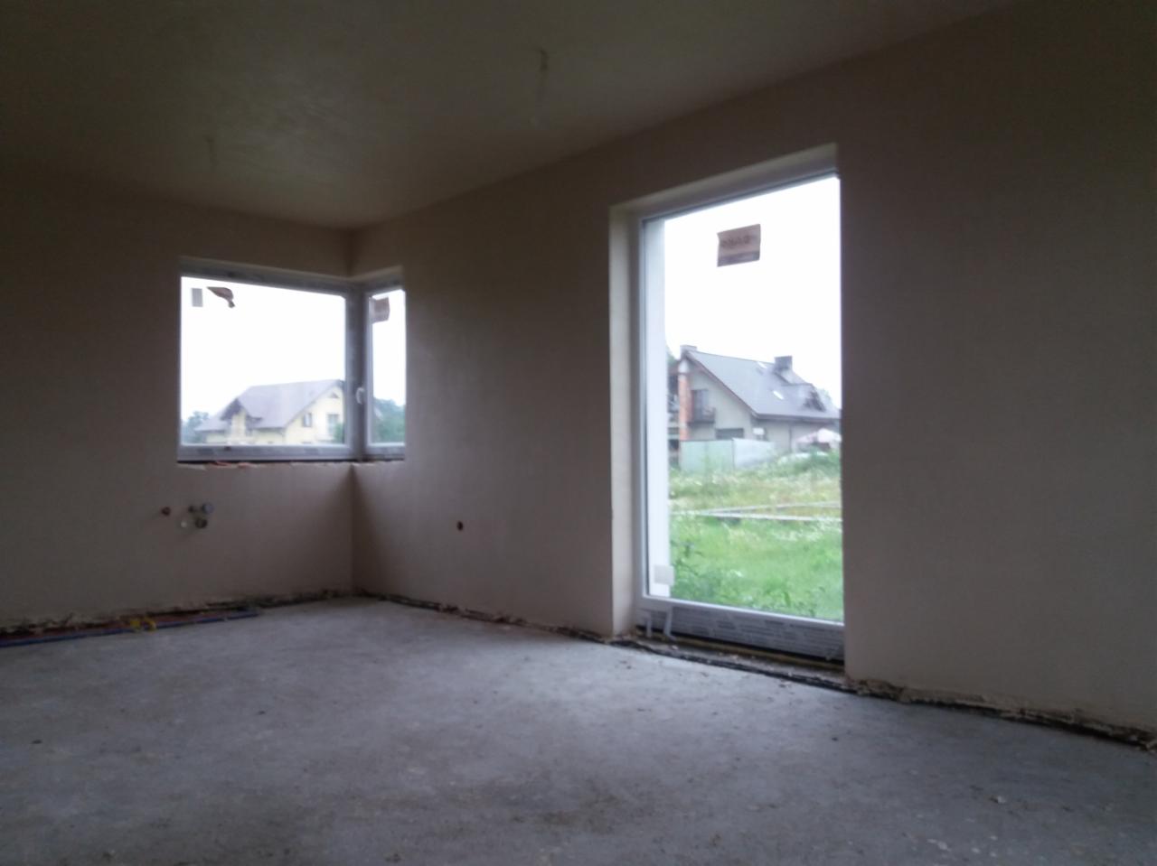 Surowe wnętrze pokoju z oknami, ściany po nałożeniu tynku maszynowego, widoczne wystające rury instalacji przy podłodze, posadzka betonowa przed wylewką.