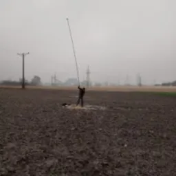 Geolog w terenie, stojący na polu z brązową ziemią, trzymający długi tyczkowy instrument pomiarowy skierowany pionowo w górę, w tle słupy energetyczne i mglista, szara pogoda.