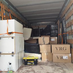 PL - Przeprowadzki Paweł Leonowicz - Wnętrze busa wypełnione kartonami VRG, meblami zabezpieczonymi pasami i folią. Widoczna skrzynka narzędziowa Stanley i butelka z płynem. Transport towarów.