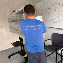PL - Przeprowadzki Paweł Leonowicz - Pracownik w niebieskiej koszulce z logo firmy przeprowadzkowej transportuje zapakowany monitor na wózku w biurze. Widoczna tablica z notatkami.