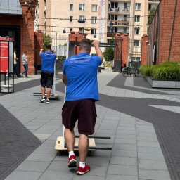 PL - Przeprowadzki Paweł Leonowicz - Dwóch mężczyzn w niebieskich koszulkach z logo firmy transportowej przesuwa metalową konstrukcję na wózku po brukowanym placu w otoczeniu budynków.