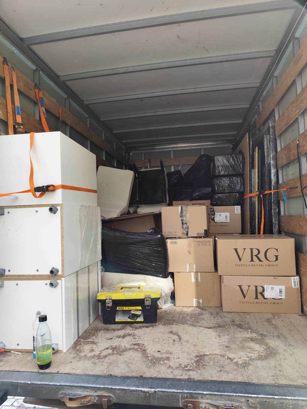 Wnętrze busa wypełnione kartonami VRG, meblami zabezpieczonymi pasami i folią. Widoczna skrzynka narzędziowa Stanley i butelka z płynem. Transport towarów.
