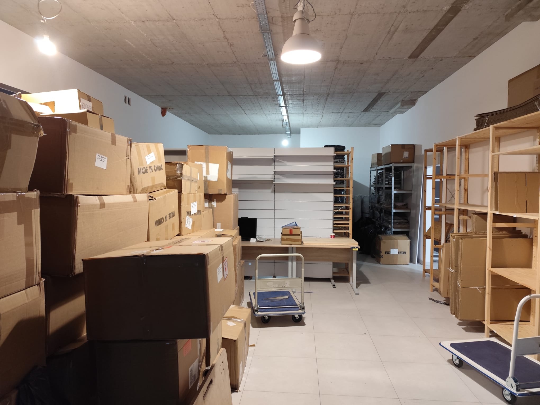 Magazyn z kartonami, regałami i wózkiem transportowym. Kartony z napisem 'Made in China'.