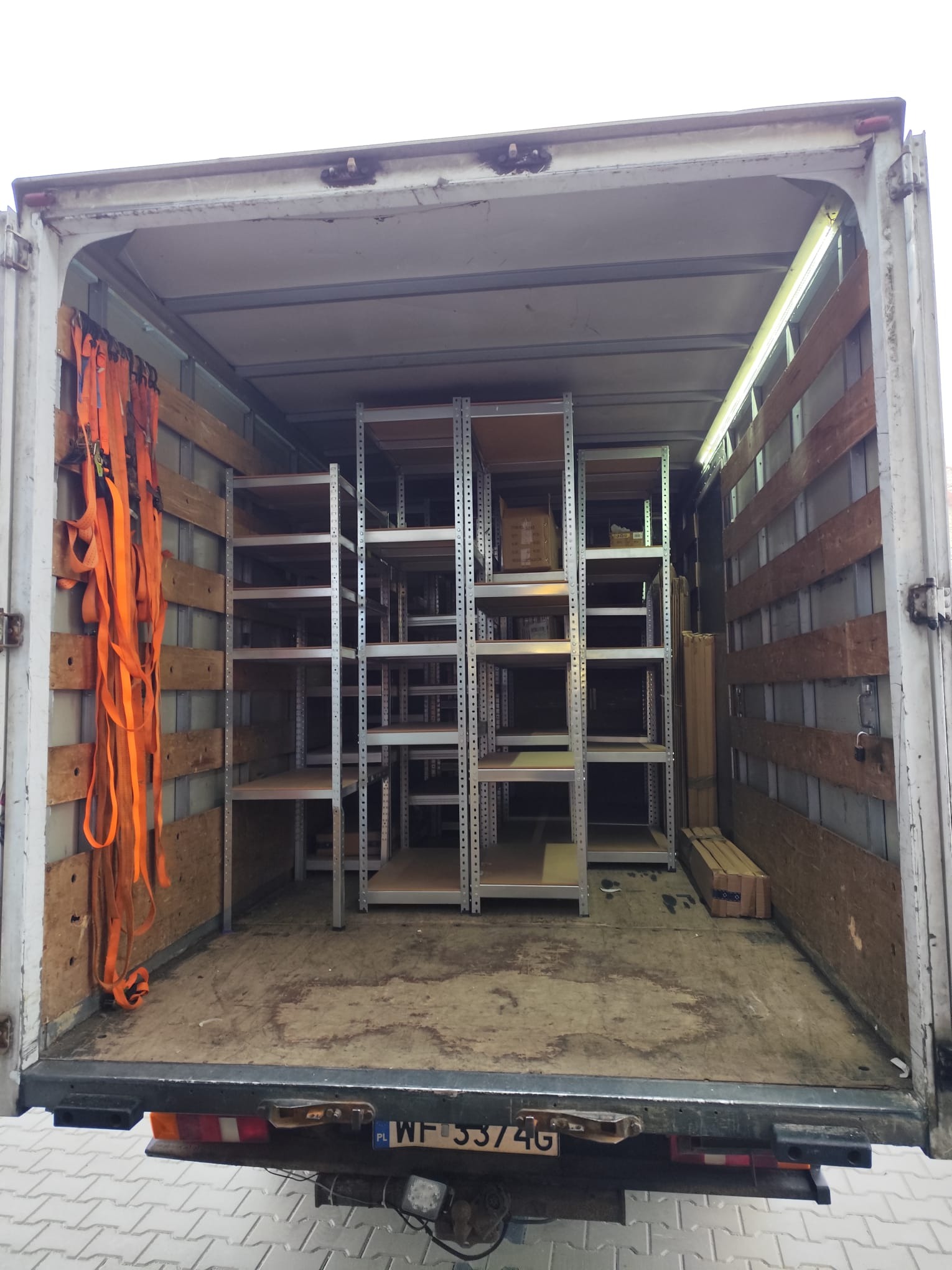 Wnętrze paki busa wypełnione metalowymi regałami i zabezpieczone pomarańczowymi pasami transportowymi, gotowe do przewozu. Widok z tyłu pojazdu.