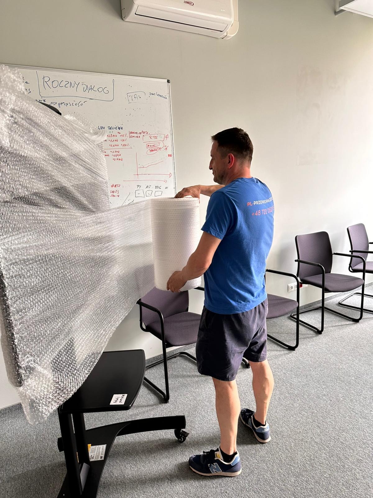 Pracownik firmy przeprowadzkowej zabezpiecza mebel folią bąbelkową w biurze. W tle biała tablica z notatkami i krzesła. Widoczny numer telefonu firmy na koszulce.