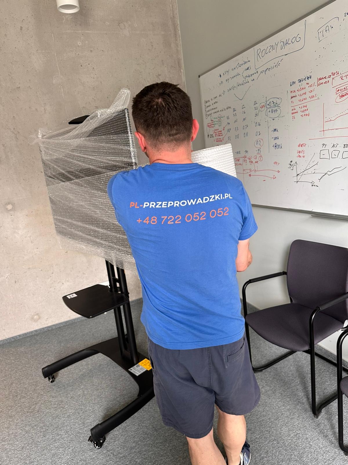 Pracownik w niebieskiej koszulce z logo firmy przeprowadzkowej transportuje zapakowany monitor na wózku w biurze. Widoczna tablica z notatkami.