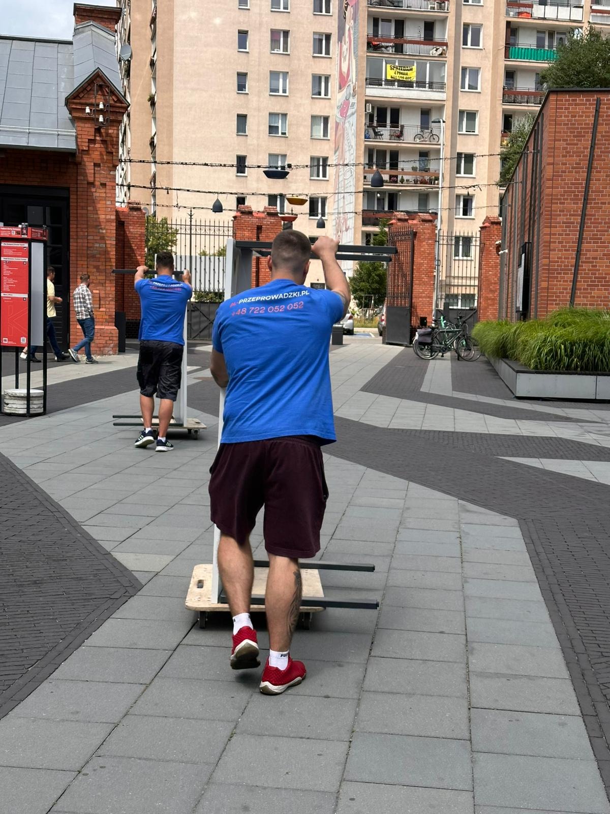 Dwóch mężczyzn w niebieskich koszulkach z logo firmy transportowej przesuwa metalową konstrukcję na wózku po brukowanym placu w otoczeniu budynków.