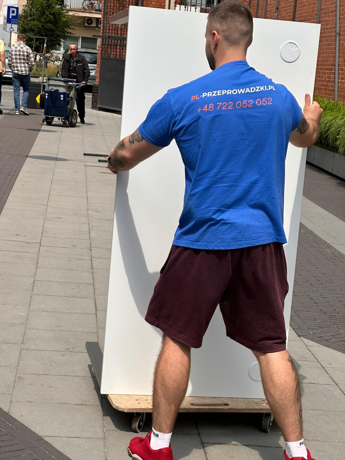 Mężczyzna w niebieskiej koszulce z logo firmy przeprowadzkowej transportuje białe drzwi na wózku na kółkach po chodniku. Widoczny fragment ulicy i budynki w tle.