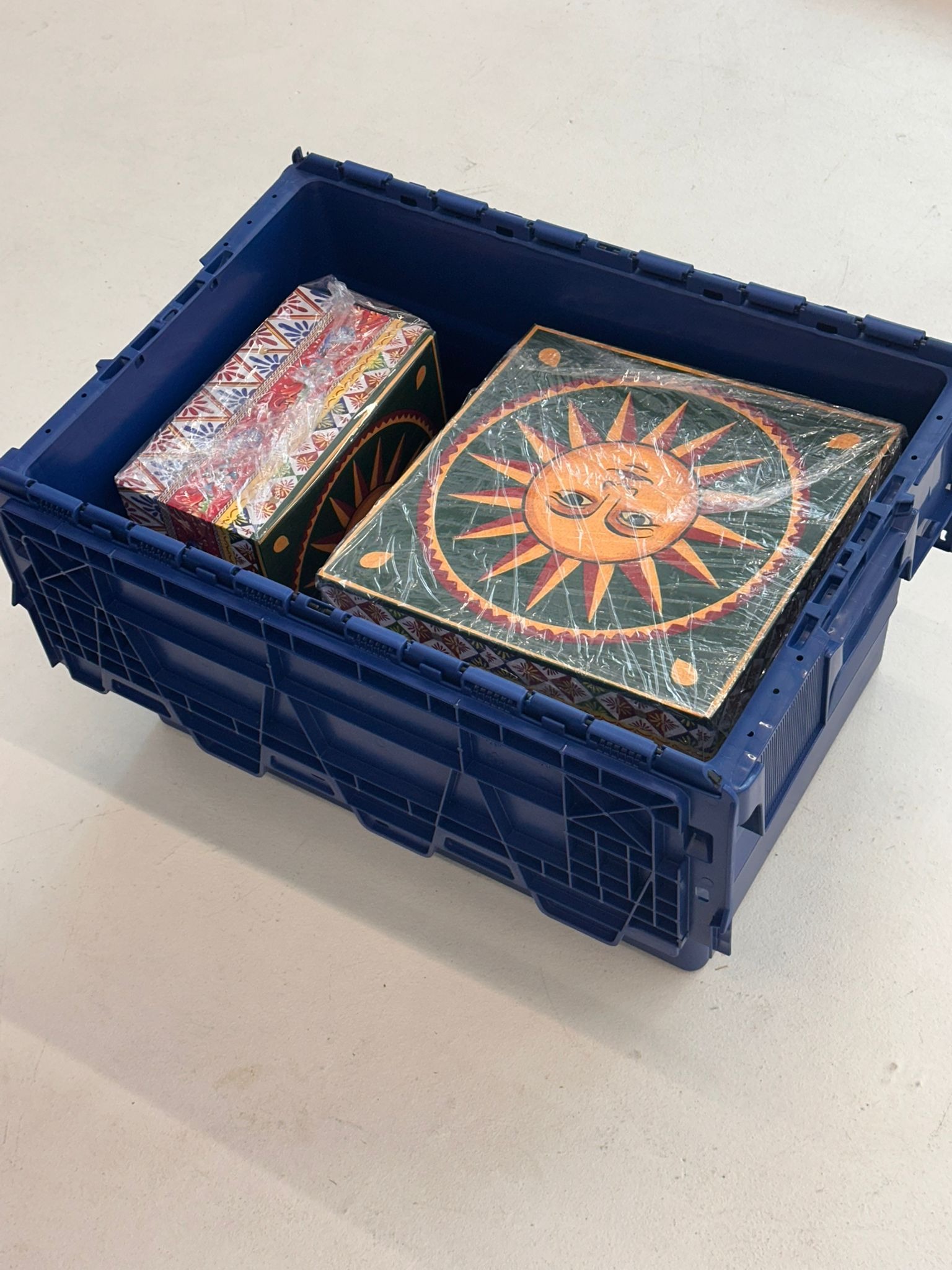 Niebieski pojemnik z dwoma zapakowanymi przedmiotami: pudełko z wzorem i płaski przedmiot ze słońcem, gotowe do transportu.