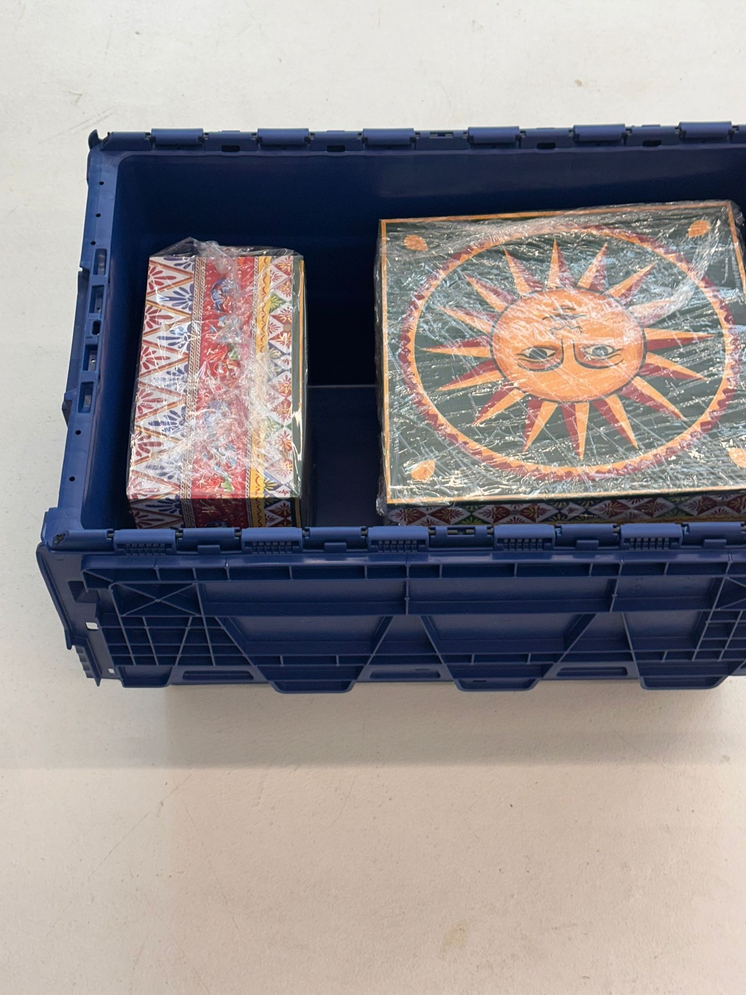 Niebieski plastikowy pojemnik z dwoma zapakowanymi przedmiotami: wąskim i wysokim oraz kwadratowym z motywem słońca, zabezpieczone folią stretch.