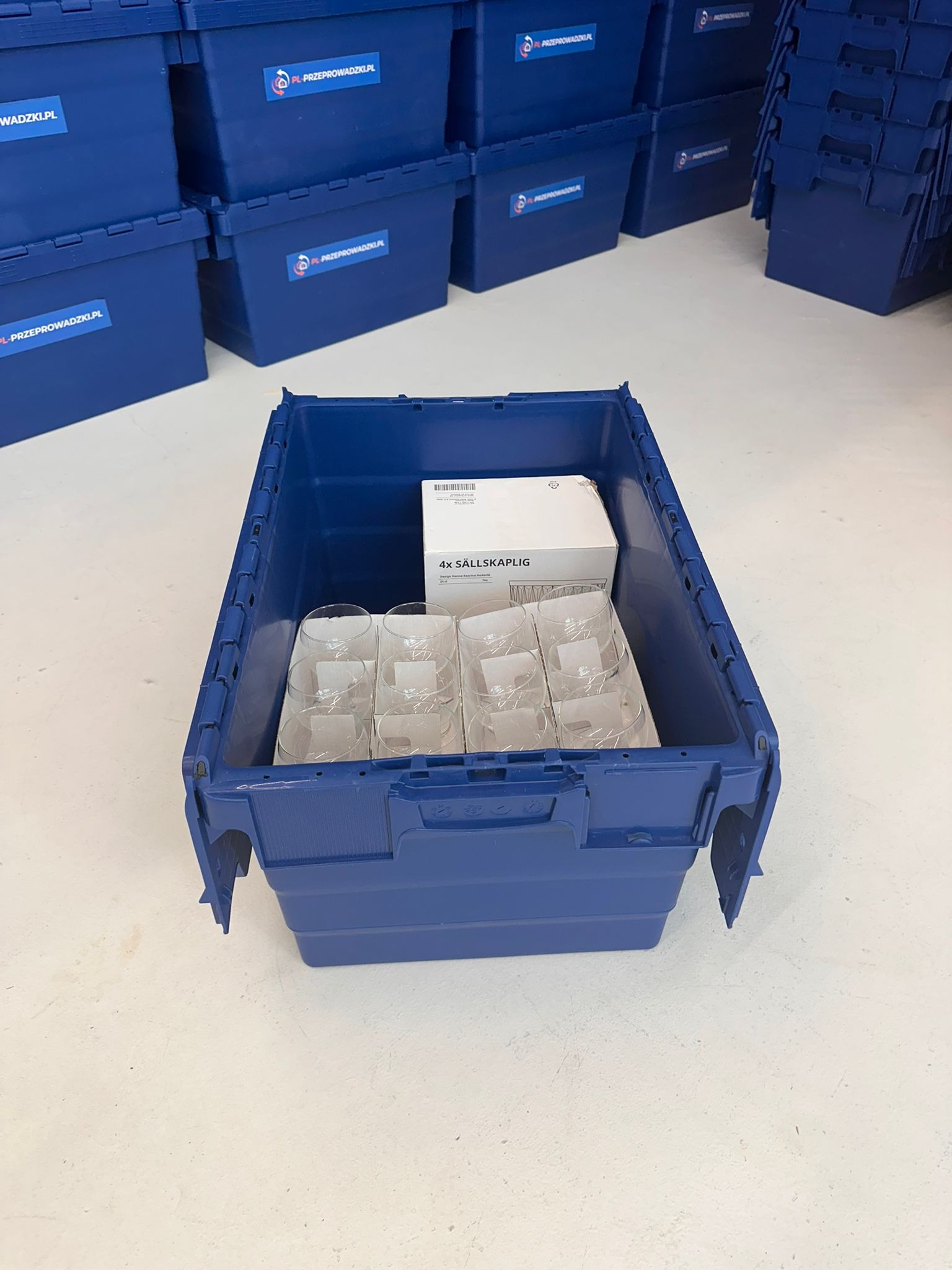 Niebieski pojemnik do przeprowadzki wypełniony szkłem i pudełkiem, otoczony stosem identycznych pojemników z logo firmy. Bezpieczne pakowanie delikatnych przedmiotów.