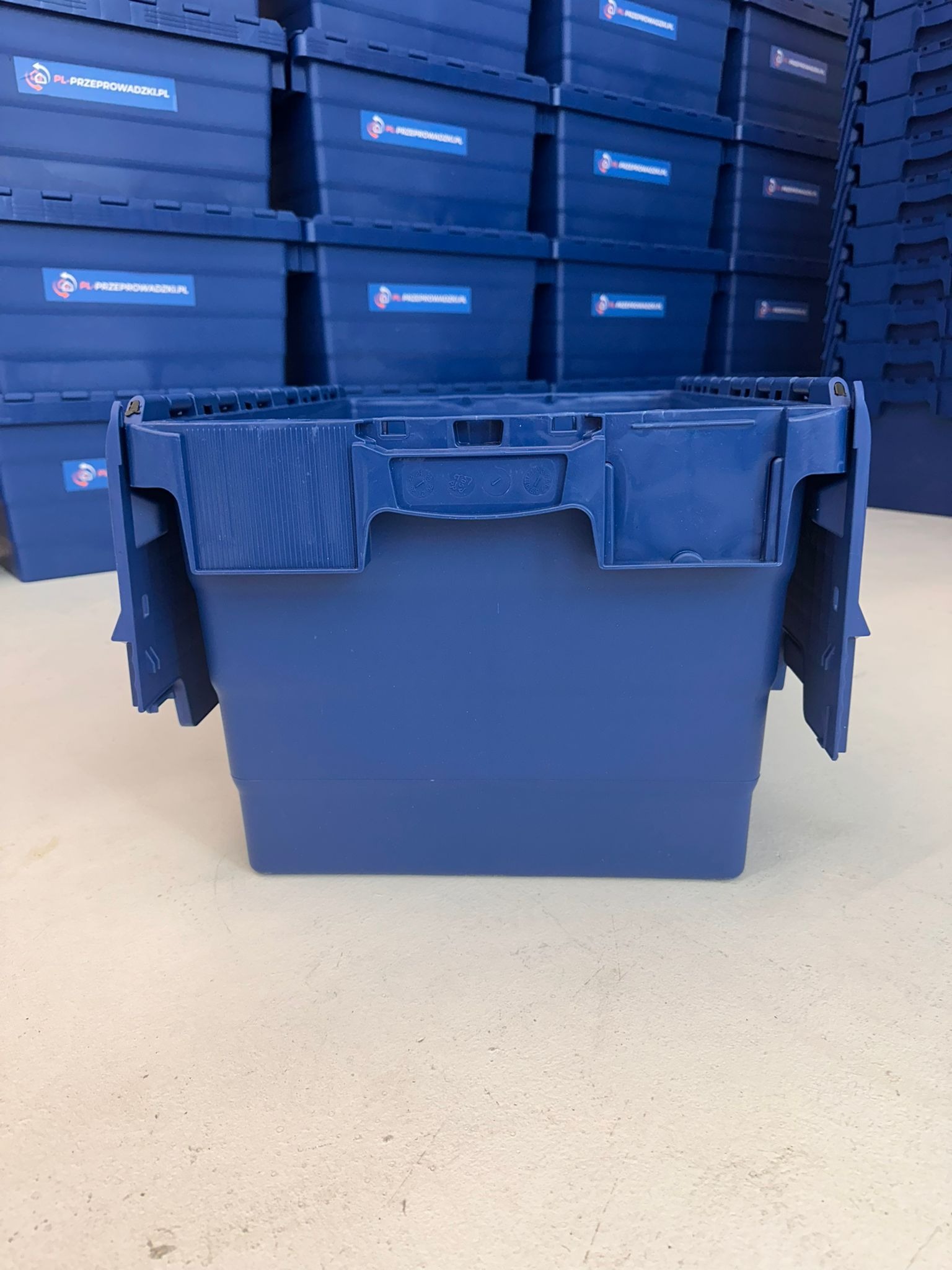 Niebieski, otwarty pojemnik z tworzywa sztucznego na tle stosu identycznych pojemników z logo firmy przeprowadzkowej. Widok z przodu, na jasnym podłożu.
