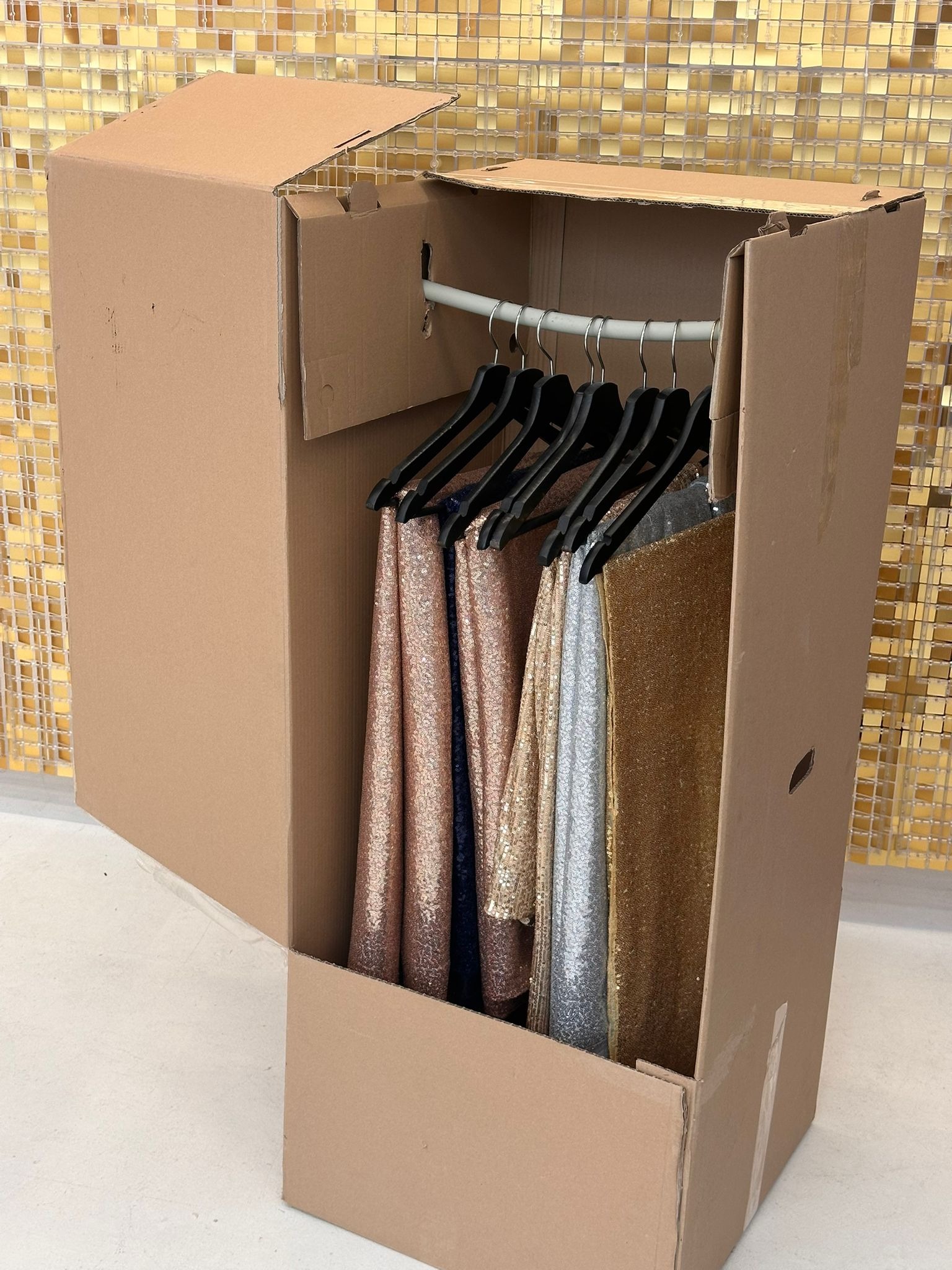 Kartonowy garderobiany kontener transportowy z błyszczącymi ubraniami na wieszakach, gotowy do przeprowadzki. Tło: złota mozaika.