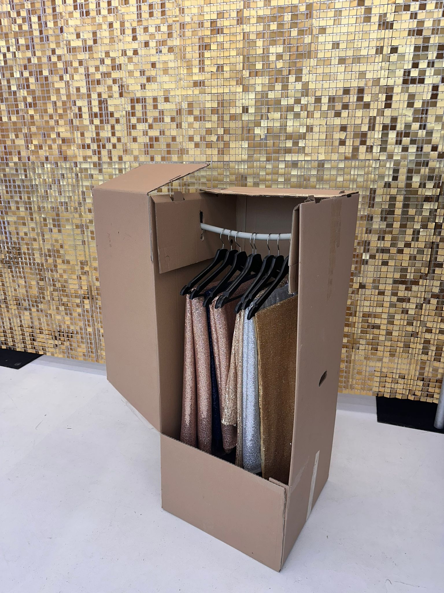 Kartonowy garderoba z błyszczącymi sukniami na wieszakach, gotowa do transportu. Złota mozaika w tle. Nietypowe rozwiązanie do bezpiecznego przewozu odzieży.