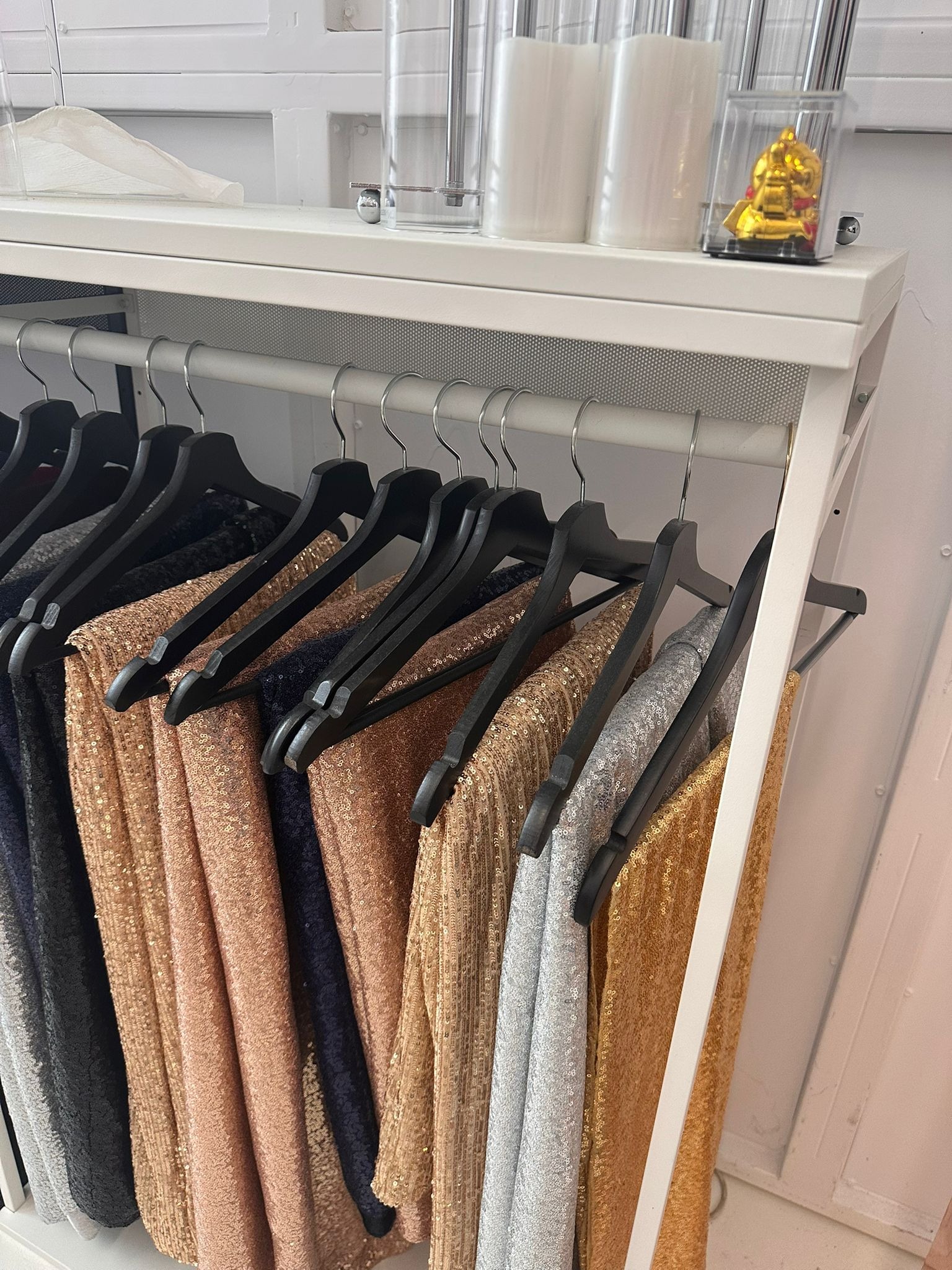 Rząd błyszczących, cekinowych tkanin w odcieniach złota, srebra i granatu, wiszących na czarnych wieszakach na tle białego regału. Dekoracje na górnej półce.