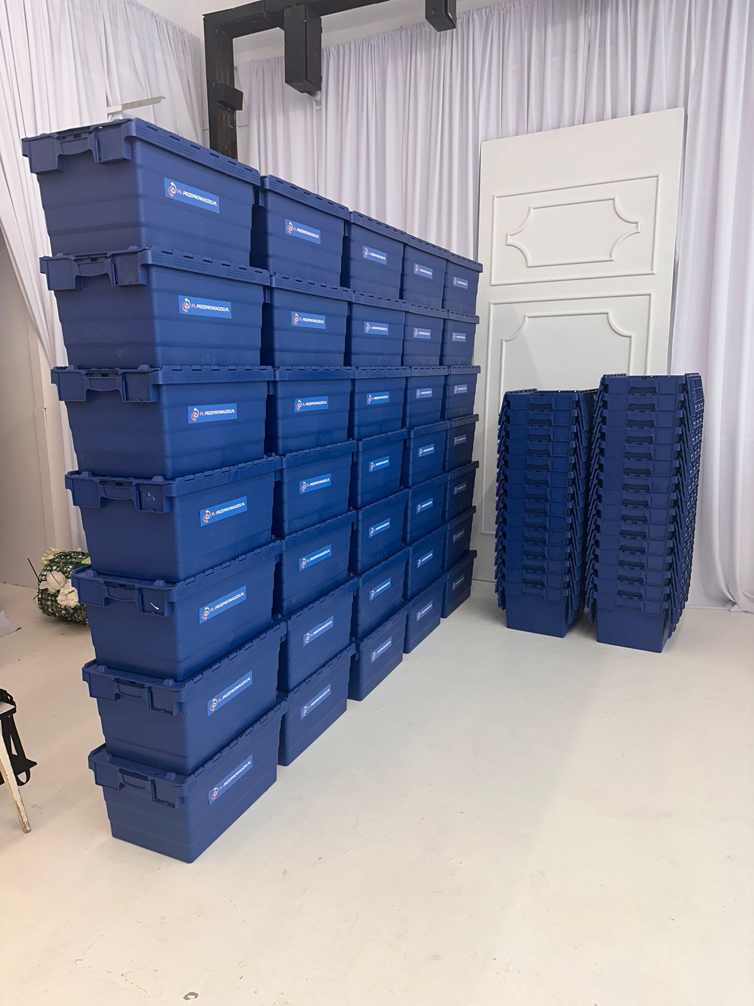Stos skrzyń do przeprowadzki z logo firmy, ustawionych w rzędach i kolumnach, obok stos pustych skrzyń, na jasnym tle z dekoracyjną ścianką.