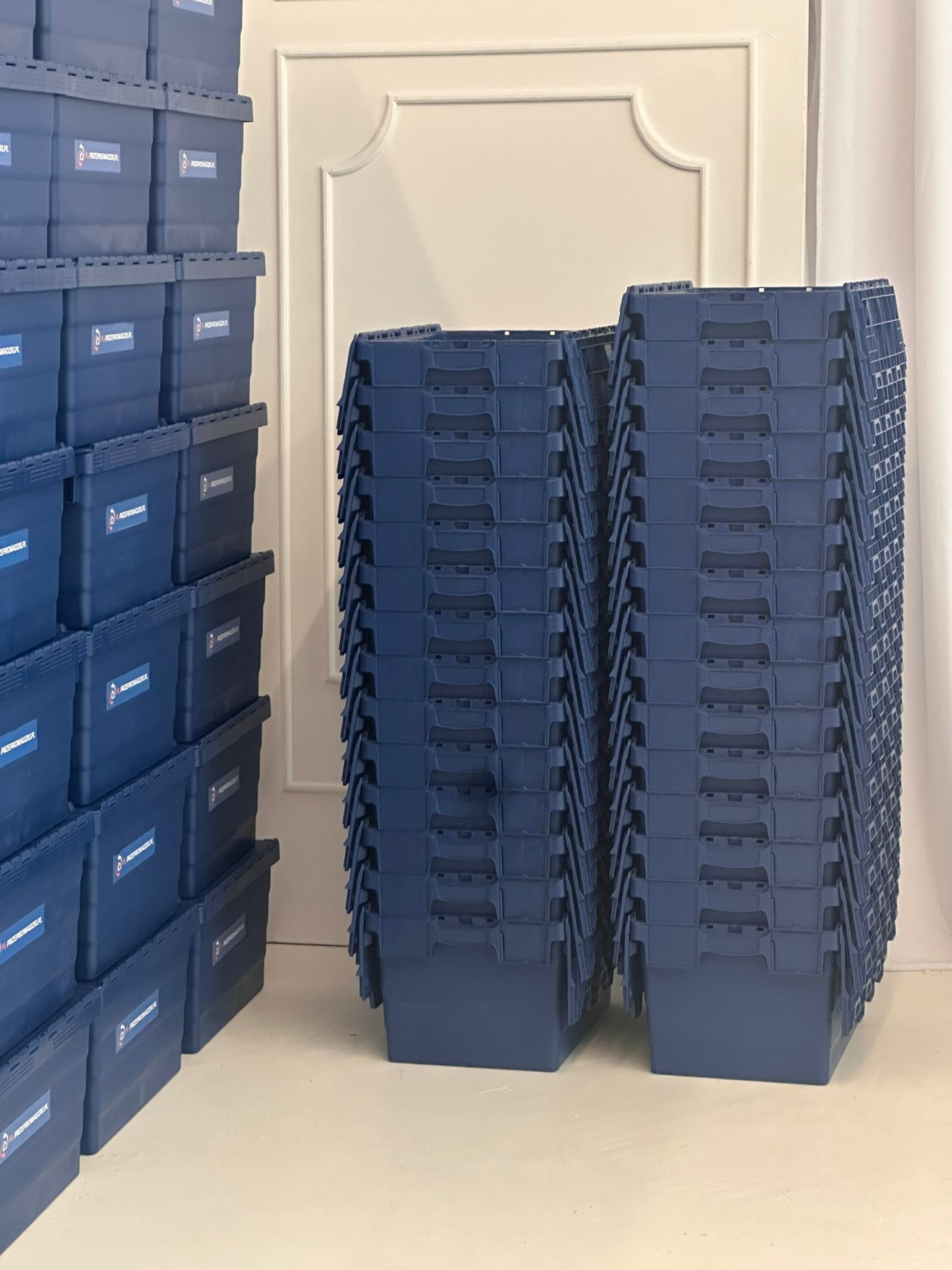 Stosy niebieskich, plastikowych pojemników do przeprowadzki, ustawione w magazynie. Widoczne logo firmy na boku pojemników. Przygotowane do transportu.