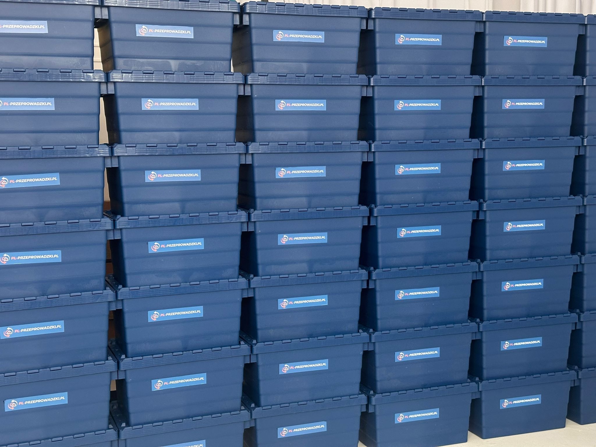 Stos niebieskich, plastikowych pojemników do przeprowadzki, z naklejkami 'PL-PRZEPROWADZKI.PL'. Pojemniki ułożone równo, jeden na drugim, tworząc uporządkowany wzór.