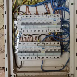 Rozdzielnica elektryczna Hager z zainstalowanymi bezpiecznikami i podłączonymi przewodami w kolorach niebieskim, żółto-zielonym i brązowym, zamontowana na ścianie z cegły i resztkami starego tynku.