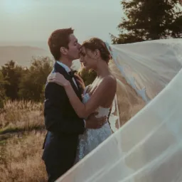 Para młoda w objęciach na tle pejzażu, panna młoda w koronkowej sukni z rozwianym welonem, pan młody w granatowym garniturze całuje ją w czoło.