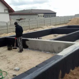 Pracownik zabezpiecza fundamenty domu przed wilgocią, używając pędzla i czarnej substancji hydroizolacyjnej, widoczny zarys ścian fundamentowych, w tle dom w trakcie budowy i ogrodzenie.