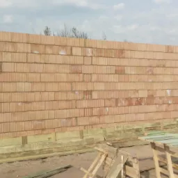 Wzniesiona ściana z czerwonej cegły ceramicznej na budowie domu, widoczne drewniane elementy szalunku u podstawy ściany, w tle fragment ogrodzenia i dachu sąsiedniego domu.