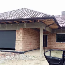 Surowy, jednopiętrowy dom z garażem w trakcie budowy, widoczne ściany z cegły, betonowe nadproża, zamontowane okno i częściowo otwarta brama garażowa, zarys samochodu z otwartymi drzwiami w prawym...