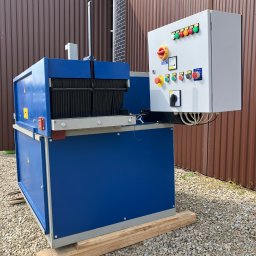 LEASING S.O.S. - Piła wielotarczowa w tartaku, z niebieską obudową i panelem sterowania. Stoi na drewnianych belkach na żwirowym podłożu, tło z brązowej blachy falistej.