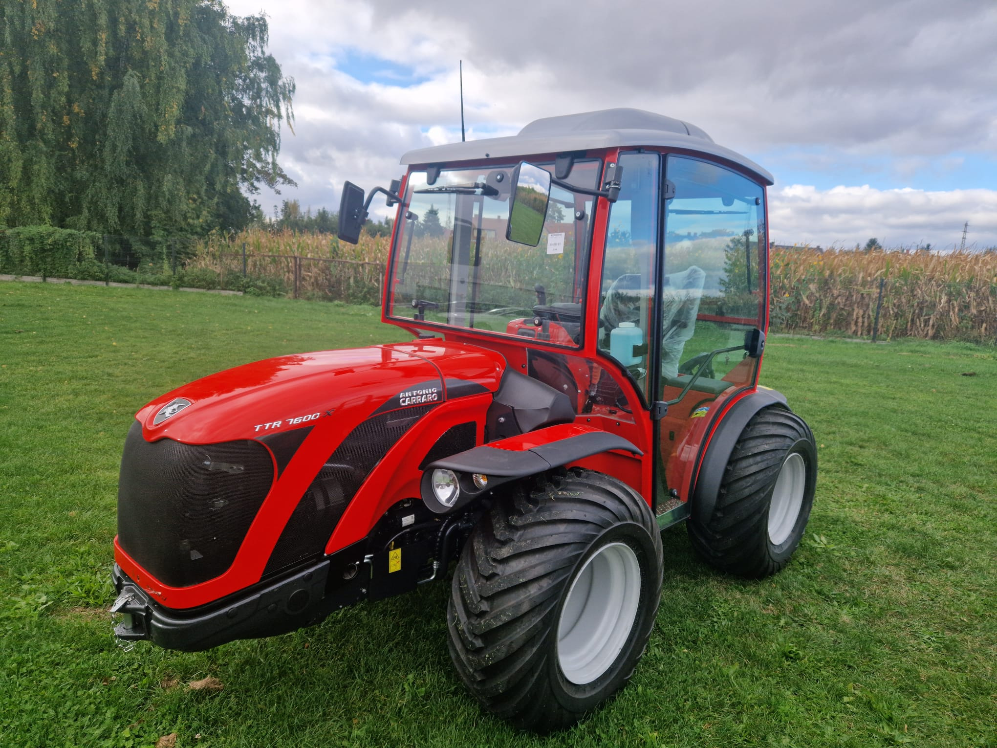 Czerwony ciągnik Antonio Carraro TTR 7600 X z kabiną na zielonej trawie, widok z przodu, w tle drzewa i pole kukurydzy, pochmurne niebo.