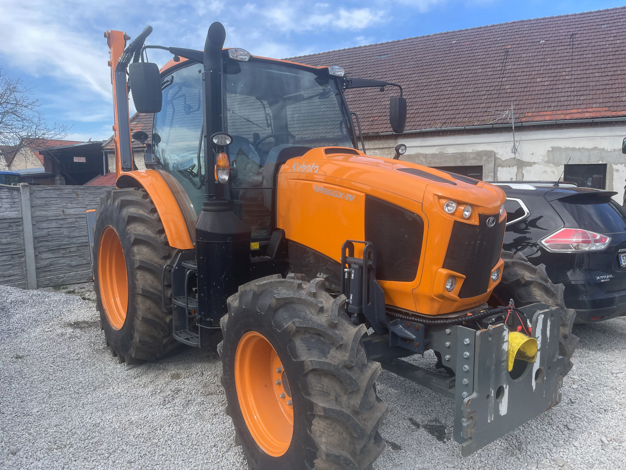 Pomarańczowy ciągnik Kubota M135GX-IV na żwirowym podłożu, z fragmentem ciemnego samochodu w tle. Widoczne detale maszyny i opony.