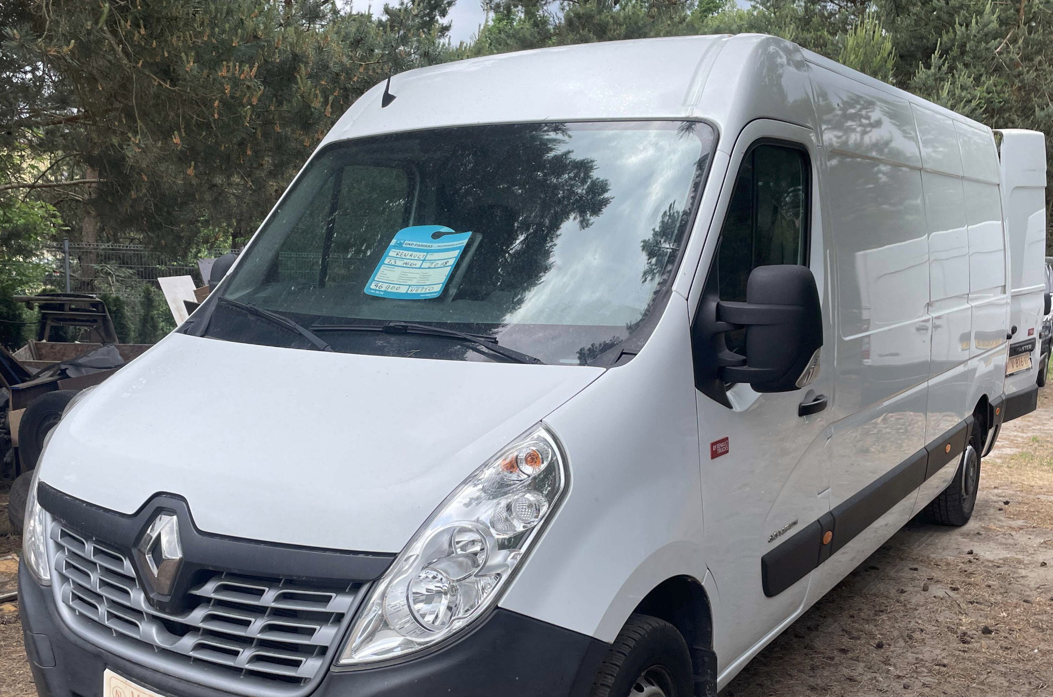 Biały Renault Master, częściowo widoczny z przodu, z kartką informacyjną na przedniej szybie, stojący na szutrowym podłożu na tle drzew.