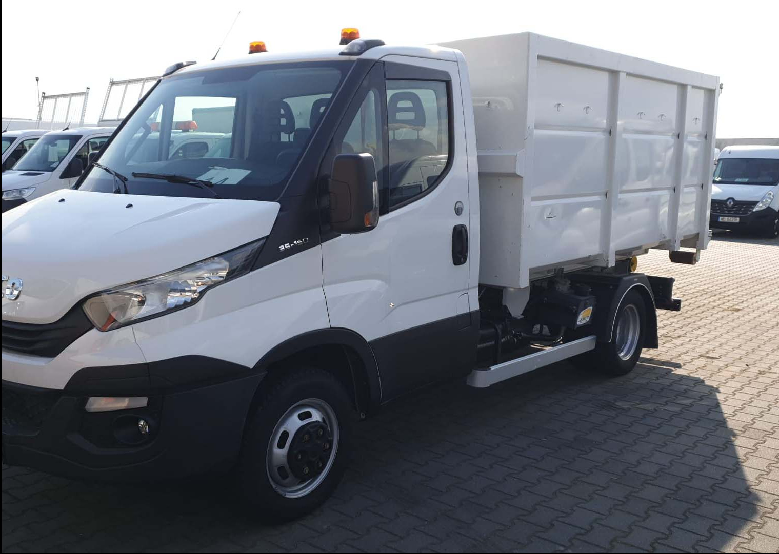 Biała mini-ciężarówka IVECO z zabudową skrzyniową, zaparkowana na placu. Widoczne detale: pomarańczowe światła ostrzegawcze na dachu, oznaczenie modelu 35-150.