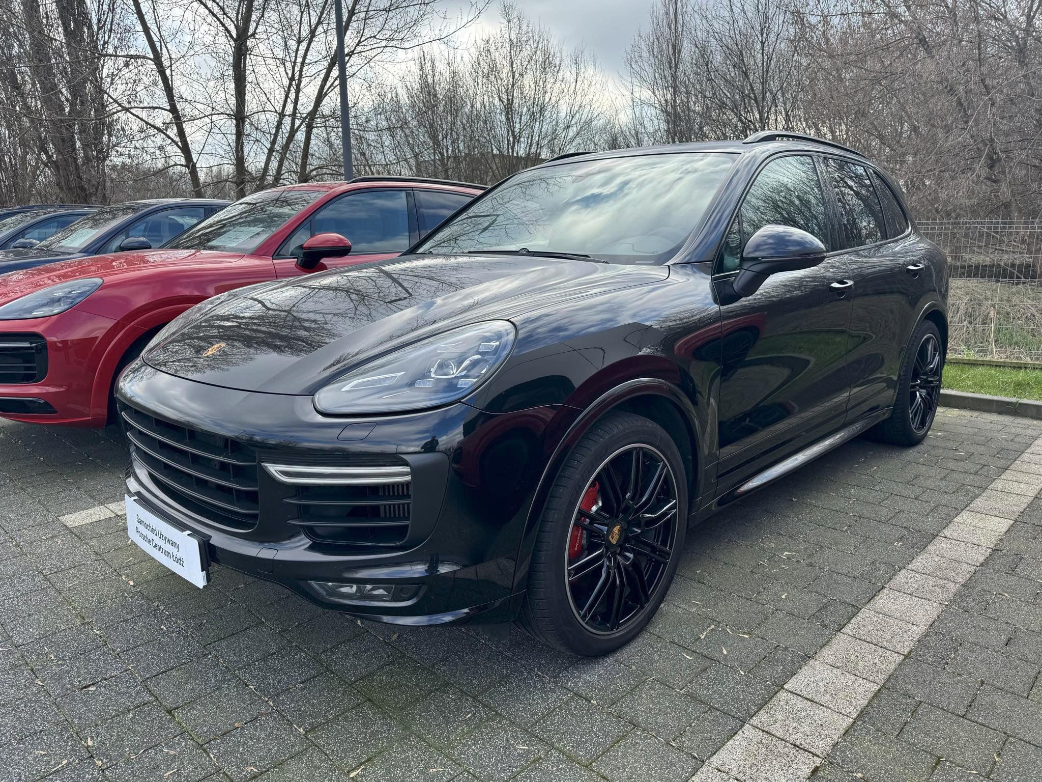 Czarny Porsche Cayenne na tle innych aut. Tabliczka z napisem 'Samochód Używany Porsche Centrum Łódź' na przednim zderzaku. Prezentacja oferty leasingu.