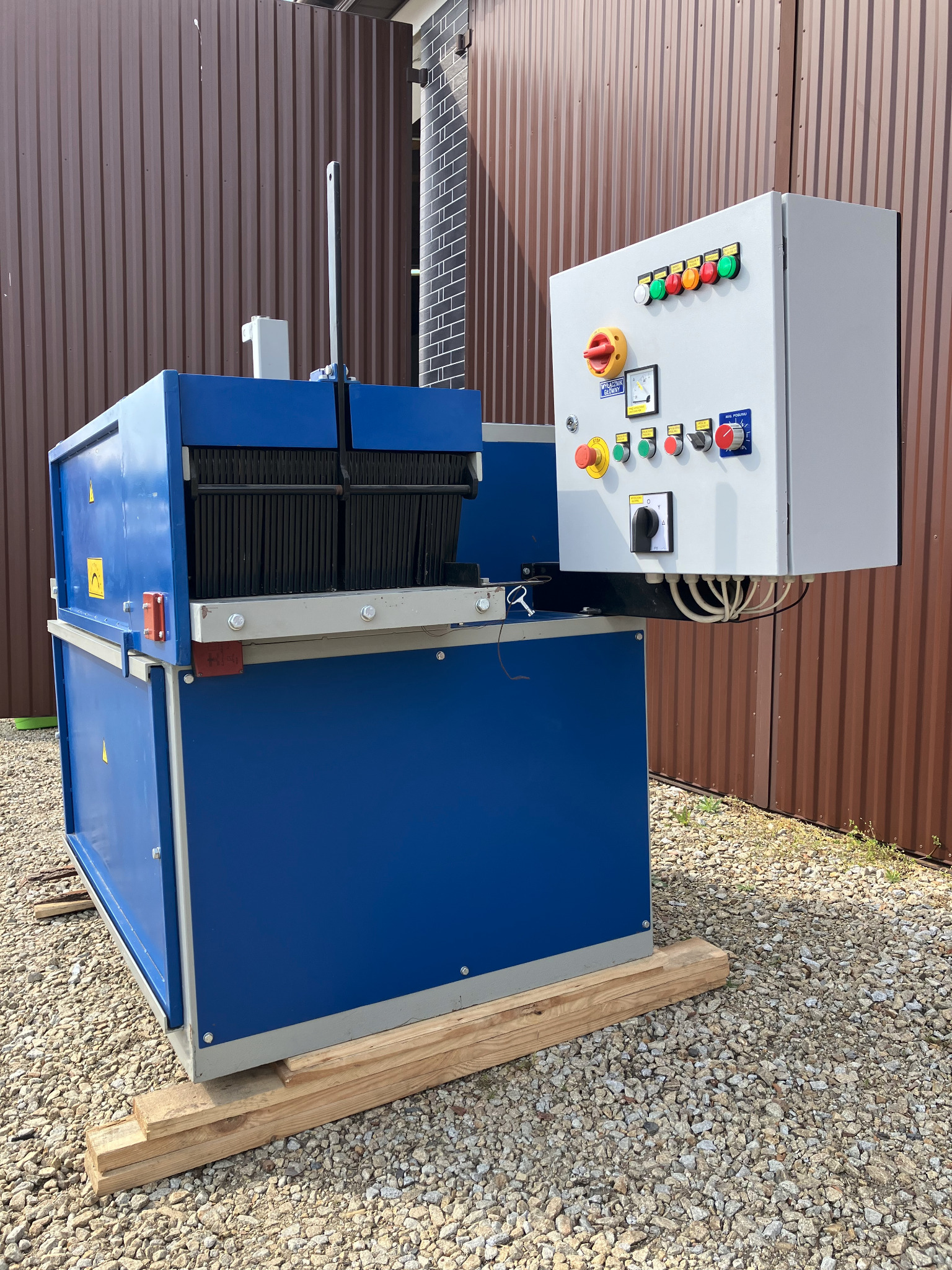 Piła wielotarczowa w tartaku, z niebieską obudową i panelem sterowania. Stoi na drewnianych belkach na żwirowym podłożu, tło z brązowej blachy falistej.