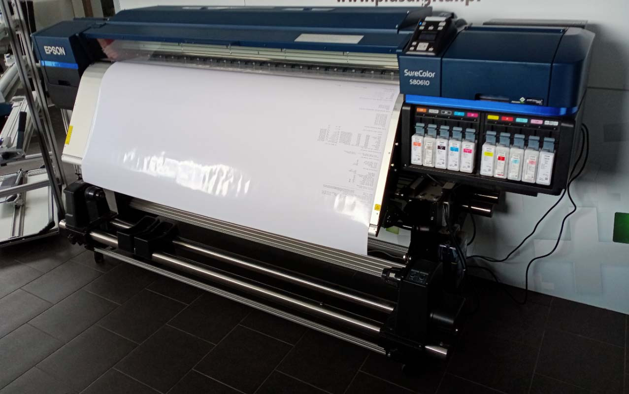 Szerokoformatowy ploter Epson SureColor S80610 z pełnym zestawem tuszy, drukujący na białym papierze z widocznymi danymi. Urządzenie stoi na ciemnej podłodze.