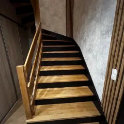 Drewniane schody z czarnymi podstopnicami, oświetlone naturalnym światłem, prowadzące do góry. Ściana wykończona panelami dekoracyjnymi.