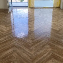 Ukończona podłoga z płytek imitujących drewno, ułożonych we wzór jodełki francuskiej. Widoczne zabezpieczenia folią malarską przy oknie i ścianie.