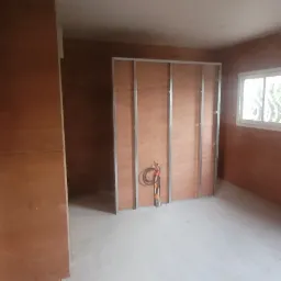 Pok&oacute;j w trakcie remontu z widocznymi drewnianymi panelami na ścianach, metalową konstrukcją ścianki działowej i surowym betonowym podłożem.