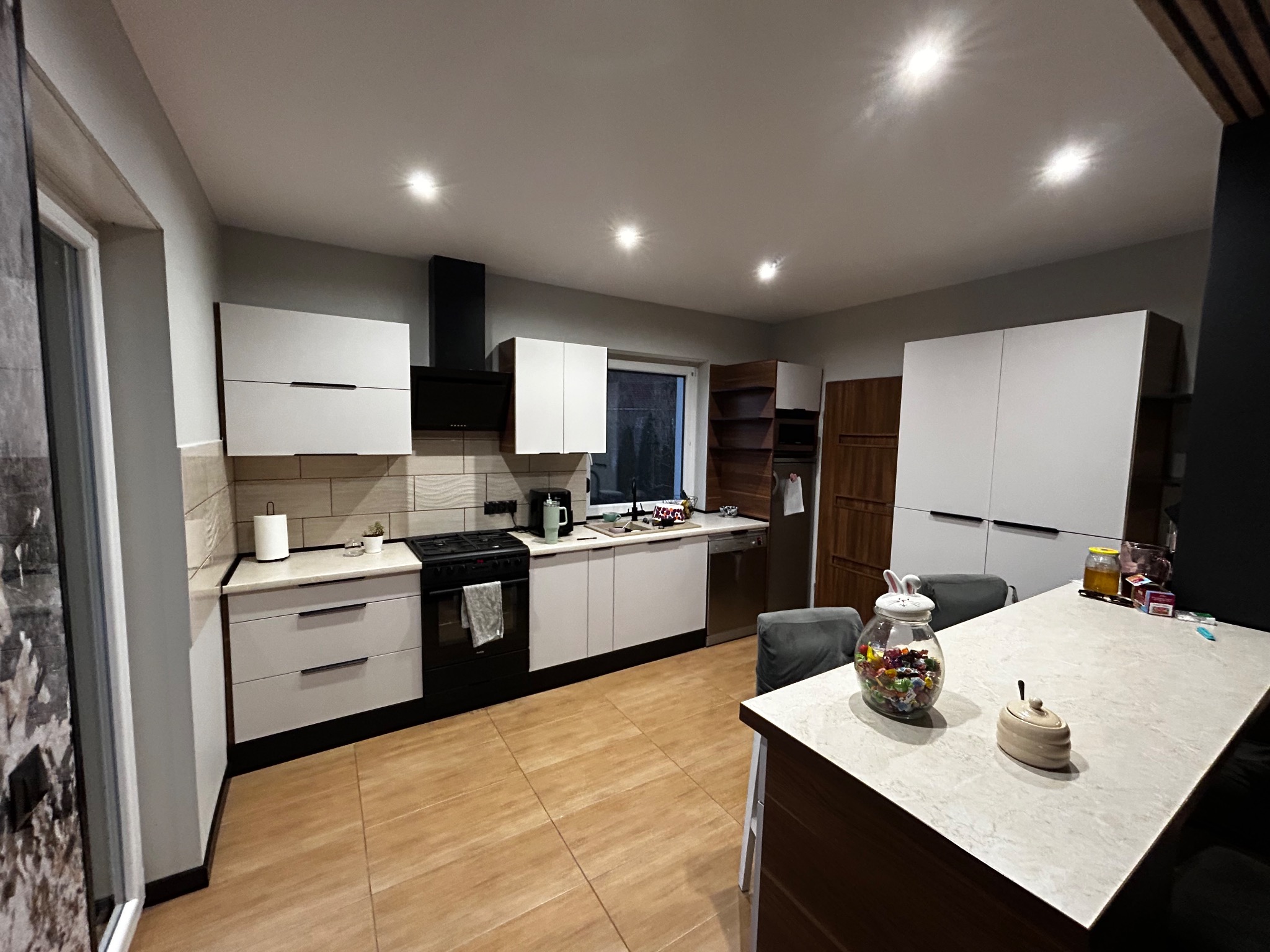 Nowoczesna kuchnia z białymi szafkami i drewnianą podłogą. Na wyspie kuchennej słoik ze słodyczami. Widoczne brązowe regały i czarny okap.