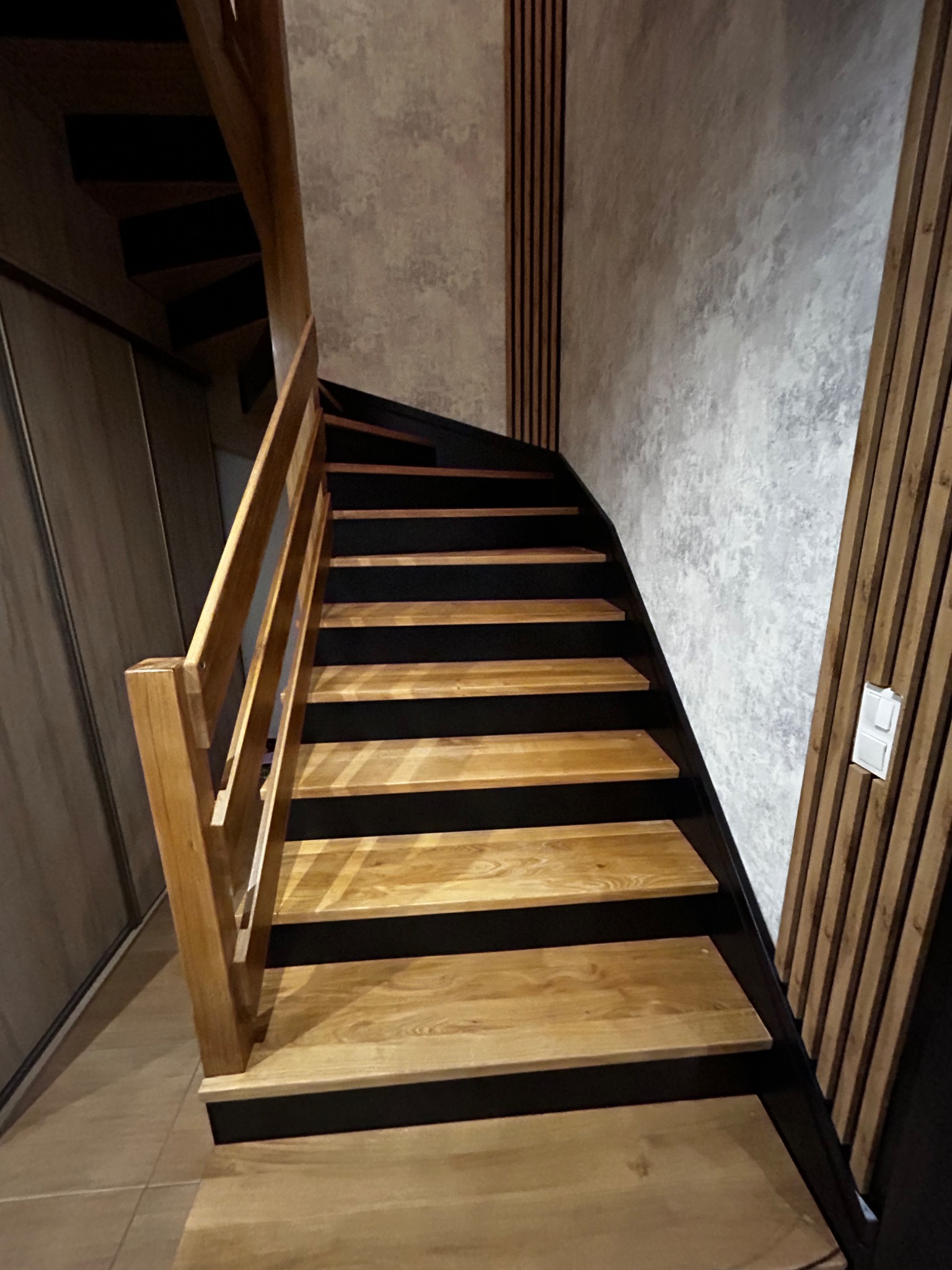 Drewniane schody z czarnymi podstopnicami, oświetlone naturalnym światłem, prowadzące do góry. Ściana wykończona panelami dekoracyjnymi.