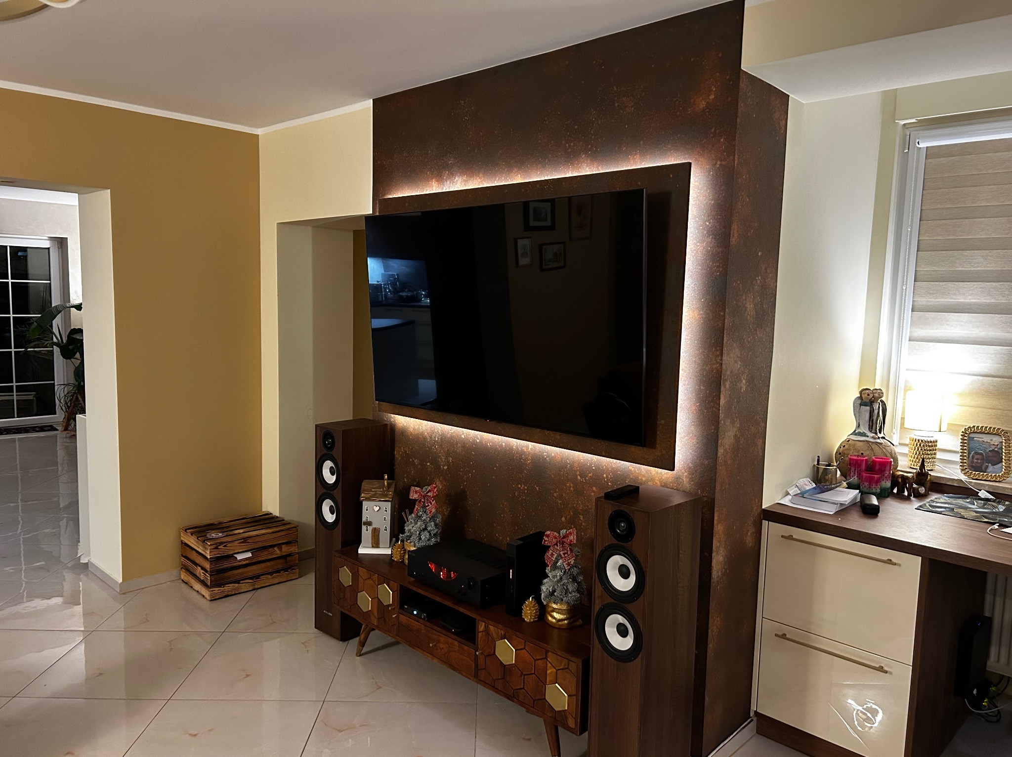 Salon z telewizorem na ścianie z efektem rdzy, podświetlonym LED. Drewniana szafka RTV i głośniki po bokach. Na komodzie dekoracje i lampka. Całość w ciepłych barwach.