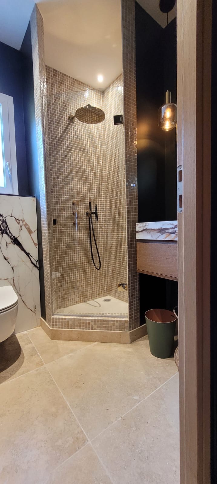 Nowoczesna kabina prysznicowa z mozaiką w beżach, czarna armatura, marmurowa umywalka i beżowa podłoga w eleganckiej łazience z ciemnymi ścianami.