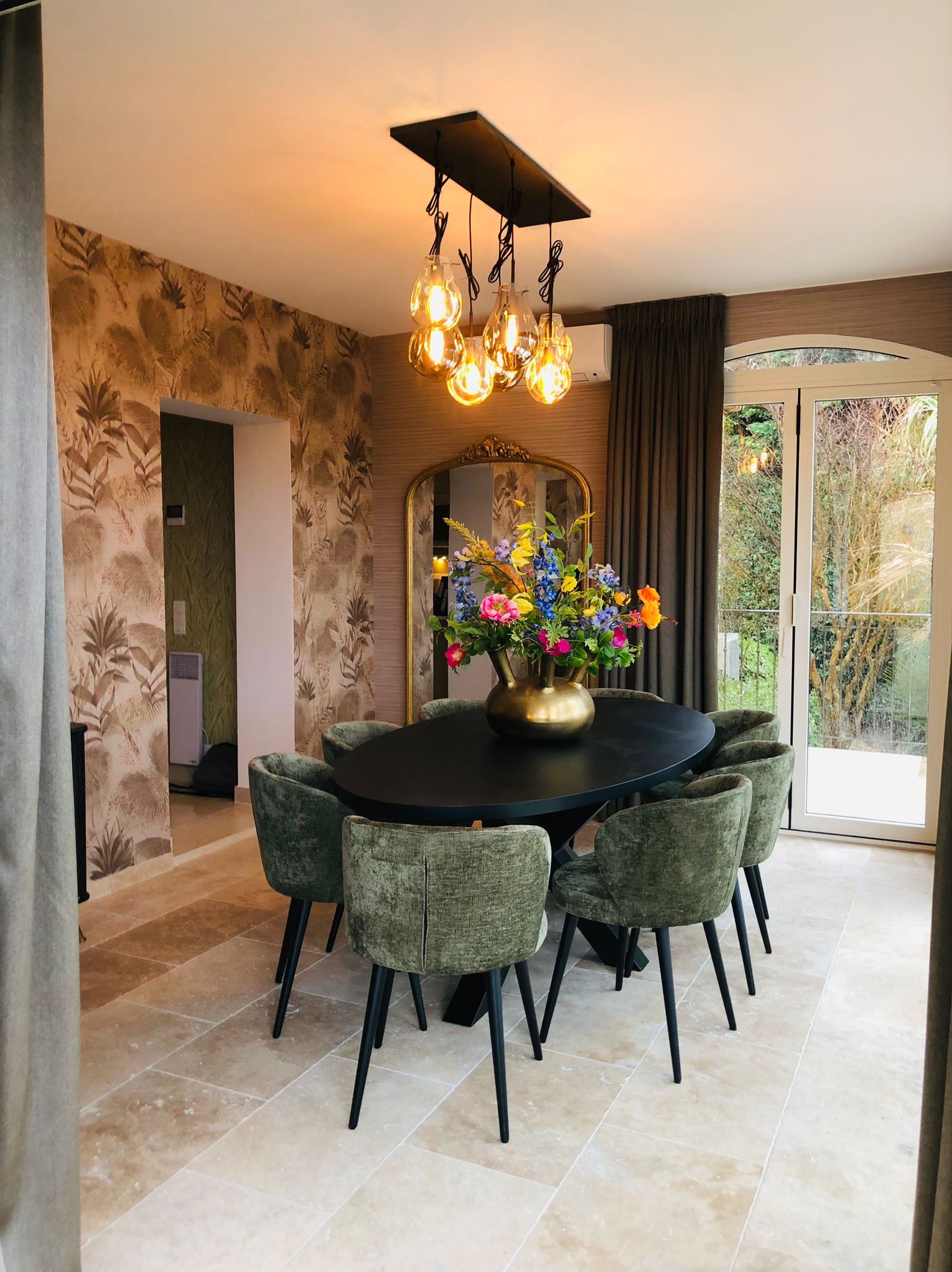 Stylowa jadalnia: owalny czarny stół otoczony zielonymi krzesłami, nad nim designerska lampa z żarówkami. W tle tapeta w roślinny wzór i lustro w złotej ramie.