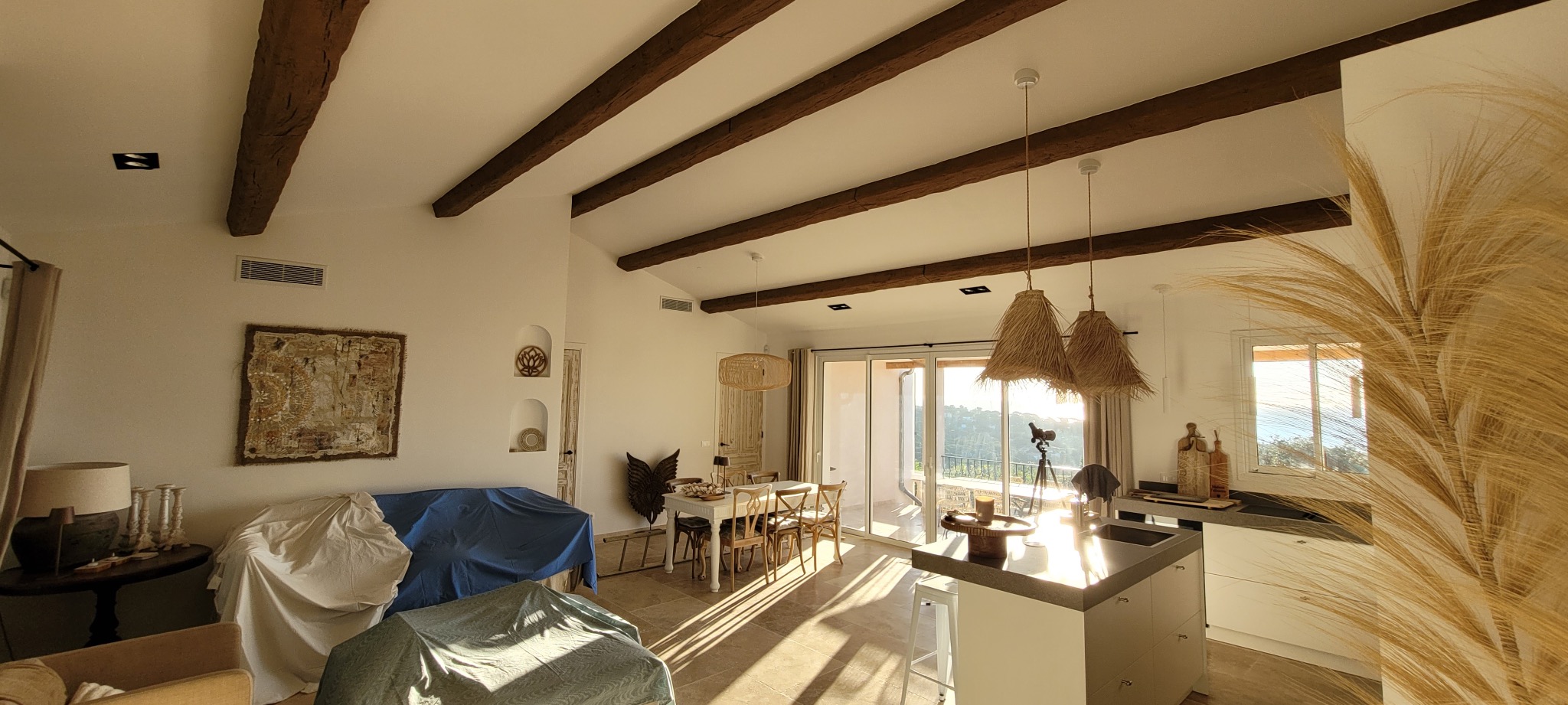 Jasne wnętrze z drewnianymi belkami, widok na krajobraz przez okno. Część mebli przykryta płachtami, sugerując prace wykończeniowe lub przygotowanie do nich.