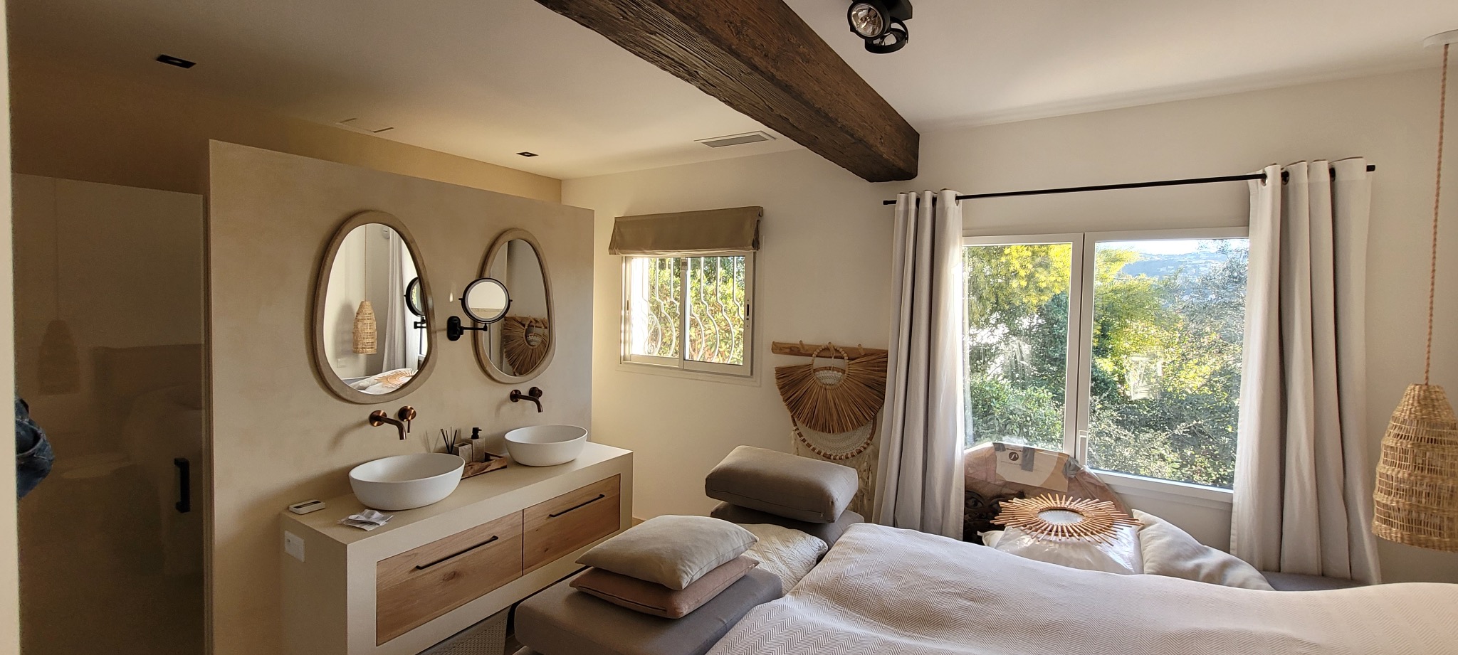 Wykończona łazienka w stylu boho: owalne lustra z drewnianymi ramami, białe umywalki nablatowe i beżowe ściany z dekoracyjną fakturą. Widoczne okno z widokiem na zieleń.