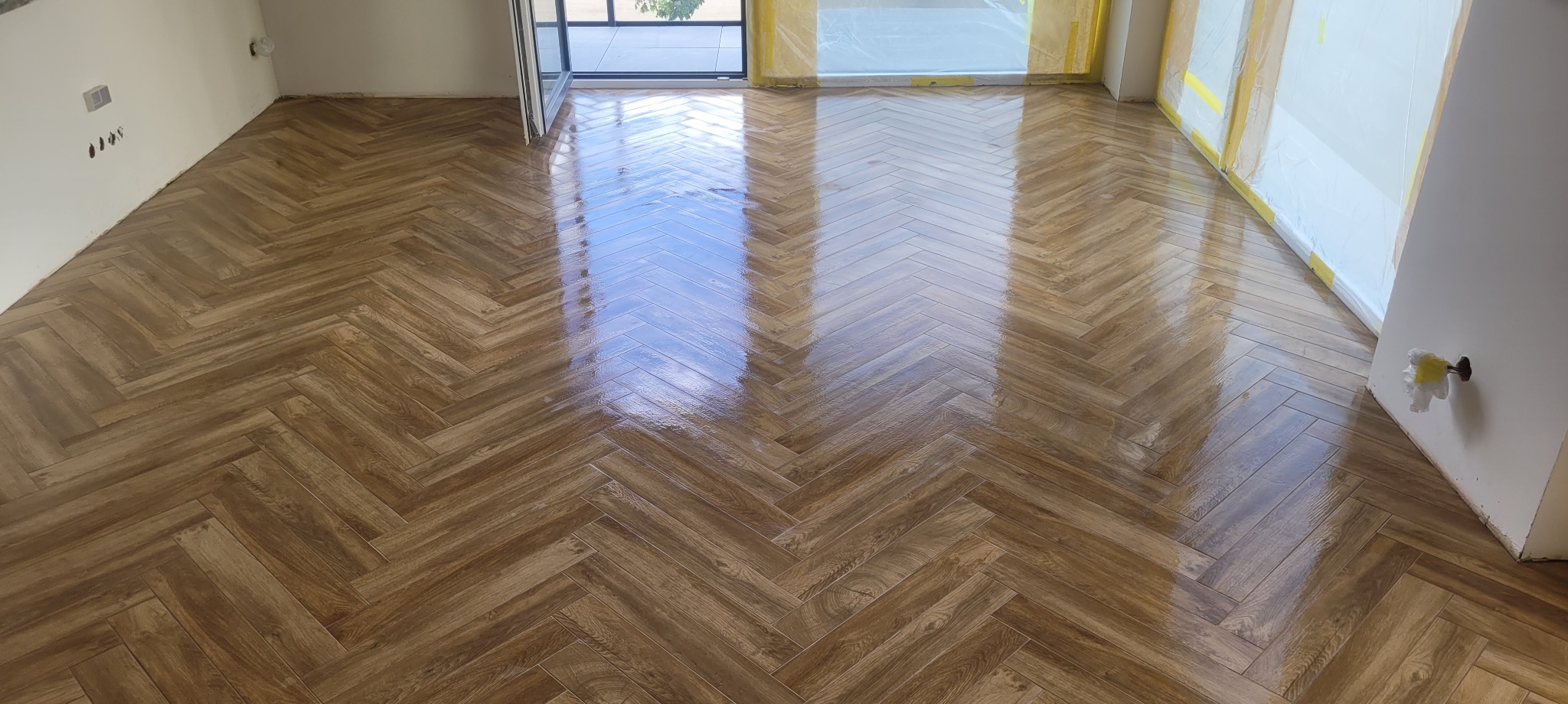 Ukończona podłoga z płytek imitujących drewno, ułożonych we wzór jodełki francuskiej. Widoczne zabezpieczenia folią malarską przy oknie i ścianie.