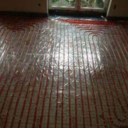 Czerwone rury ogrzewania podłogowego ułożone na siatce w pomieszczeniu przed zalaniem wylewką, widoczne okno i fragment ściany.