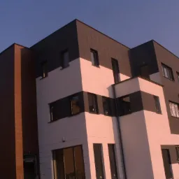 Nowoczesny budynek z elewacją w odcieniach bieli i grafitu, z elementami ceglanymi. Widoczne okna i balkon, oświetlone popołudniowym słońcem.