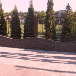 Ozdobne, kute ogrodzenie z ceglaną podmurówką, widziane z poziomu chodnika. W tle zieleń i budynek.